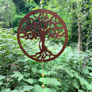 May include: A copper-colored metal tree of life wind chime with a circular design and a chain of amber-colored beads hanging from the bottom. The wind chime is hanging from a tree branch in a forest setting.