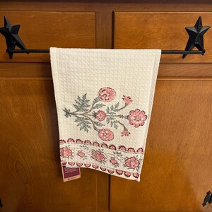 May include: A white waffle-weave kitchen towel with a floral design in pink and gray hangs from a black metal bar. The bar is supported by star-shaped brackets. The towel features a repeating floral border.