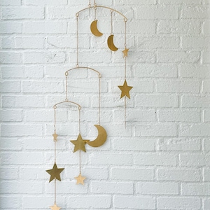 May include: A gold-colored mobile featuring crescent moons and stars suspended from arched frames. The celestial-themed nursery decor hangs against a white brick wall, creating a whimsical and decorative accent.