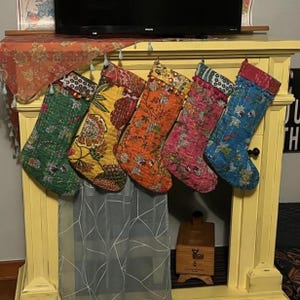 May include: Five colorful Christmas stockings hang from a yellow mantel. Each stocking features a unique floral pattern in shades of green, yellow, orange, pink, and blue. The stockings are quilted and have decorative trim.