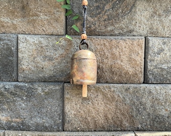 May include: A rustic, weathered brass bell with a cylindrical wooden clapper suspended from a dark, knotted cord. The cord features two small, light-colored wooden beads above the bell, which has a rounded top and a slightly flared base.