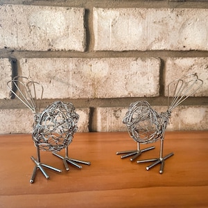 May include: Two bird figurines crafted from silver-toned wire. Each bird features a rounded, open-work body made of coiled wire, a fan-shaped tail, and thin, straight wire legs with three-pronged feet. The figures are positioned side by side.