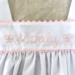 Puede incluir: Un vestido blanco de bebé con un ribete rosa festoneado y el nombre "Blakeley" bordado en hilo rosa con lazos rosas a cada lado.