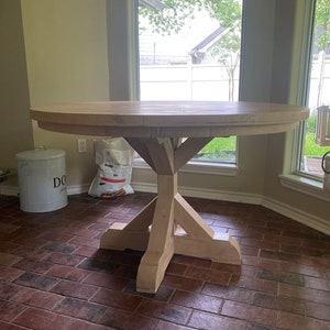 May include: A round wooden table with a pedestal base. The table is made of light wood and has a natural finish. The table is sitting on a brick floor.