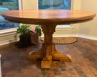 May include: A round wooden table with a natural wood finish and a pedestal base. The table has a simple, rustic design and is perfect for a dining room or kitchen.
