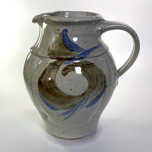 May include: A ceramic pitcher with a speckled light gray glaze, featuring a hand-painted abstract design in brown and blue brushstrokes. The vessel has a rounded body, a wide pouring spout, and a curved handle on the right side of the structure.