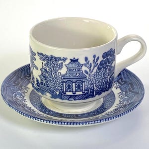 May include: A ceramic teacup and saucer set featuring a classic blue and white willow pattern. The design depicts detailed oriental-style pagodas, trees, and landscape elements printed in a deep blue ink against a creamy white background.