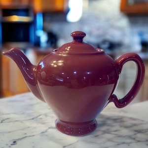 May include: A deep burgundy ceramic teapot with a rounded body and a lid. The teapot has a spout and a handle.