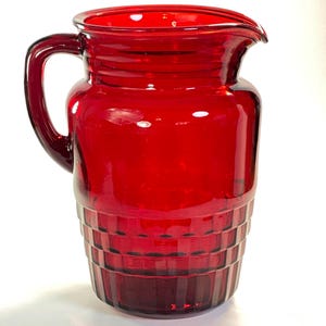 Può includere: Una caraffa di vetro rosso rubino con manico e beccuccio. La caraffa presenta un design geometrico strutturato nella metà inferiore e una parte superiore liscia e arrotondata. Il manico è curvo e attaccato al lato.