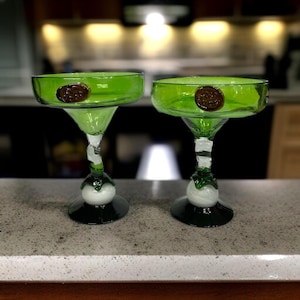 May include: Two green margarita glasses with a brown emblem on the bowl. The glasses have a unique design with a white ball and dark green base. The glasses are on a speckled countertop.