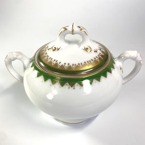 May include: A white porcelain sugar bowl with two handles and a matching lid. The piece features a decorative band of green and gold triangular patterns around the rim and the lid, with gold accents on the handle of the lid and the side handles.