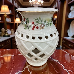 May include: Cream-colored ceramic vase with a gold rim and a lattice design. The vase features a painted floral design with red berries, green leaves, and a pinecone. The vase is a decorative home accent.