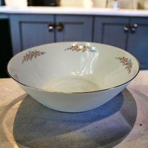 May include: A white ceramic bowl with a gold rim and a floral design. The bowl has a delicate floral pattern in shades of pink, blue, and green.