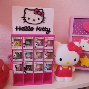 May include: A pink display case with a Hello Kitty theme, featuring a sign with the character's face and name. The case holds several collectible figures in boxes. A large Hello Kitty figure stands nearby.