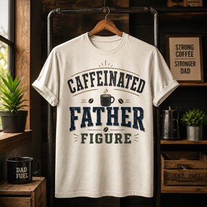 Op de afbeelding: Lichtbeige t-shirt met de woorden "CAFFEINATED FATHER FIGURE" in donkerblauwe en groene letters. Het shirt heeft een afbeelding van een koffiekopje en hangt aan een houten hanger.