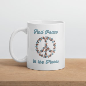 Op de afbeelding: Witte keramische mok met een blauw vredesteken gemaakt van puzzelstukjes. De tekst "Find Peace in the Pieces" staat in blauw onder het vredesteken.