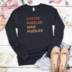May include: Charcoal grey long-sleeved top with the text "COFFEE PUZZLES WINE PUZZLES" in orange and red. The shirt is styled with denim shorts and beige flats. The background shows a floral design on a white wooden surface.