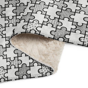 Può includere: Una coperta in pile bianco con un motivo a pezzi di puzzle bianco e nero. La coperta è piegata a metà, mostrando il morbido pile bianco all'interno.