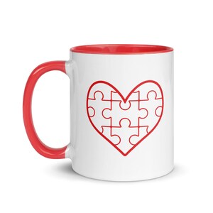 Puede incluir: Taza de cerámica blanca con borde y asa rojos. La taza presenta un contorno rojo de una forma de corazón compuesta por piezas de rompecabezas.