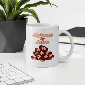 May include: White ceramic coffee mug featuring a brown and orange graphic of coffee beans and jigsaw pieces. The text "Jigsaw & Java" is printed in a retro font on the mug.