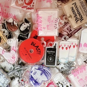 Könnte beinhalten: Eine Sammlung klarer Acryl-Anhänger mit verschiedenen Designs. Die Anhänger zeigen Illustrationen von Frauen, Texten und anderen Grafiken in Pink, Rot, Schwarz und Weiß. Einige Anhänger haben eine runde Form, andere sind rechteckig oder quadratisch.