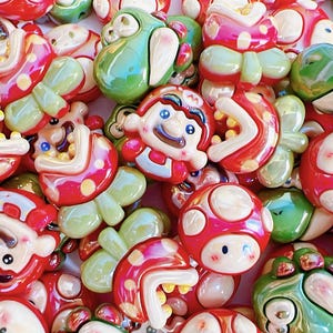 May include: A close-up of a collection of glossy resin charms in red, white, and green. The charms feature cartoon characters and mushroom designs, ideal for crafting or jewellery making. The charms vary in shape and size.