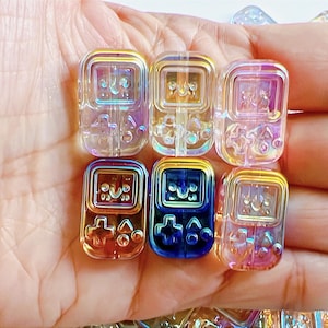 May include: Six iridescent game console-shaped beads in a hand. The beads are in a variety of colours, including yellow, blue, and purple. The beads have a glossy finish.