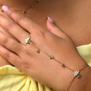 May include: A gold-toned hand chain featuring two floral-inspired motifs set with clear marquise-cut stones and small dangling beads. The piece connects a ring component to a wrist bracelet via a delicate chain adorned with evenly spaced metal beads.