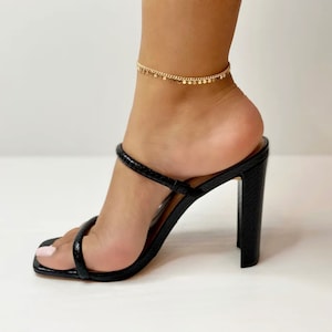Könnte beinhalten: Schwarze High-Heel-Sandalen mit quadratischer Zehenpartie und Schlangenlederstruktur. Ein zartes goldenes Fußkettchen mit kleinen kreisförmigen Anhängern schmückt den Knöchel. Die Sandalen haben einen dicken Absatz.