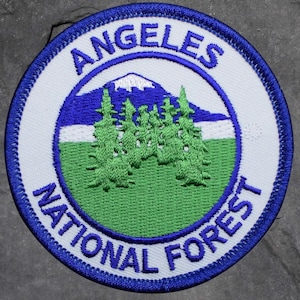 May include: Embroidered patch featuring the text "ANGELES NATIONAL FOREST" in blue, arched around a central design. The design includes a mountain range with a snow-capped peak, green trees, and a green and blue background.
