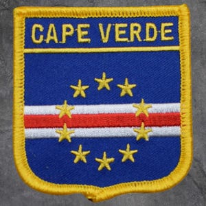 May include: A patch displaying the Cape Verde flag design. The patch has a blue background with a white, red, and white horizontal stripe. Seven yellow stars are arranged in a semi-circle. The words "CAPE VERDE" are in yellow at the top, with a yellow border.