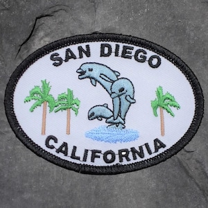 May include: Oval patch featuring the text "SAN DIEGO CALIFORNIA" in black. The patch has a white background with a design of three blue dolphins jumping over blue water, and two green palm trees.