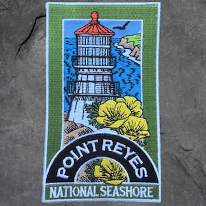 May include: Embroidered patch featuring the Point Reyes Lighthouse with a red roof, set against a blue sky and ocean. Yellow flowers and the words "POINT REYES NATIONAL SEASHORE" are also included. The patch has a green border.