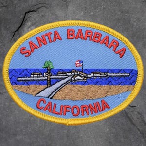 Peut inclure: Écusson brodé avec un fond bleu portant les mots "SANTA BARBARA" et "CALIFORNIA" en rouge. L'écusson représente une jetée, un palmier et l'océan. L'écusson a une bordure jaune.