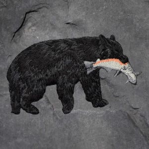 May include: Embroidered patch showing a black bear with a fish in its mouth. The fish has orange and white details. The background is a textured grey. A small bird flies above the bear.