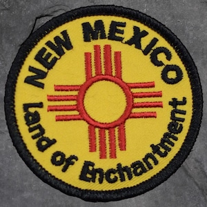 May include: Round yellow patch with black border featuring the words "NEW MEXICO Land of Enchantment" in black text. The center has a red and yellow Zia symbol.