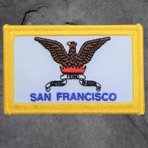 May include: Embroidered patch featuring a brown eagle with outstretched wings above a red flame. The patch has a white background with the words "SAN FRANCISCO" in blue and a yellow border. The word "FERRO" is on a banner.