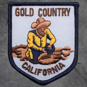 May include: Embroidered patch in a shield shape, depicting a gold miner in yellow and blue clothing, sitting on a brown log. The text "GOLD COUNTRY CALIFORNIA" is arched above and below the miner. The patch has a white background and a navy blue border.