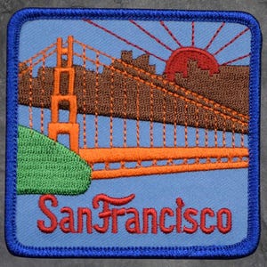 May include: Embroidered patch featuring the Golden Gate Bridge in orange and brown, with a red sun setting behind the city skyline. The word "San Francisco" is stitched in red across the bottom. The patch has a blue border and a light blue background.