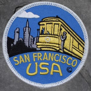 May include: A round patch with a blue background featuring a yellow cable car and San Francisco skyline. The text "SAN FRANCISCO USA" is in yellow. The patch has a white stitched border.