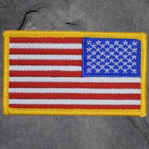 May include: Embroidered rectangular patch depicting the American flag. The patch features red and white stripes, a blue canton with white stars, and a yellow border. The design is a detailed representation of the United States flag.