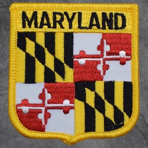May include: Embroidered patch featuring the Maryland state flag design. The patch has a yellow border and the word "MARYLAND" in black block letters at the top. The flag design includes black and gold diagonal stripes and a red and white cross.
