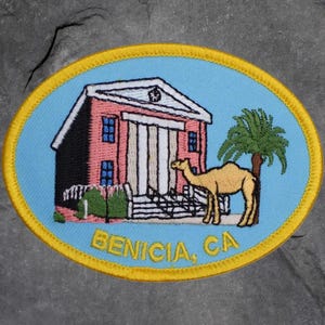May include: Embroidered patch with a light blue background and a yellow border. The patch features a red building with white columns, a camel, and a palm tree. The text "BENICIA, CA" is embroidered in yellow.