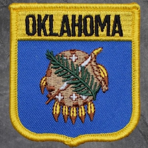 May include: Embroidered patch with the word "OKLAHOMA" in black on a yellow border. The patch has a blue background with a detailed emblem of a bison, olive branch, and Native American symbols.