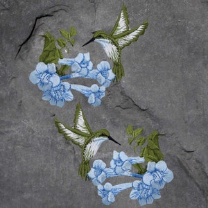 May include: Two embroidered hummingbirds with green bodies and white wings are perched on blue flowers with green leaves. The hummingbirds are facing opposite directions. The embroidery is on a gray background.
