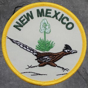 Peut inclure: Écusson brodé circulaire avec l'inscription "NEW MEXICO" en vert. Il représente un grand géocoucou et une plante de yucca sur fond crème, avec une bordure jaune.