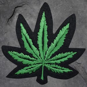 May include: Embroidered patch with a green cannabis leaf design and detailed stitching. The leaf is set against a black background. This patch is suitable for adding to clothing or accessories, offering a distinctive look.