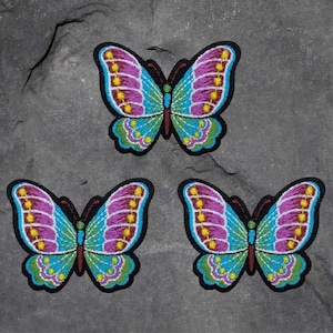 May include: Three embroidered butterfly patches with a black border. The butterfly wings are a combination of purple, turquoise, and yellow. The butterfly's body is green and brown. The patches are on a gray, textured surface.