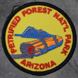 以下が含まれることがあります： 黄色い背景と黒い縁取りの丸い刺繍パッチ。パッチには赤い文字で「PETRIFIED FOREST NATL PARK ARIZONA」と書かれています。青とオレンジの丸太のイラストが中央にあります。