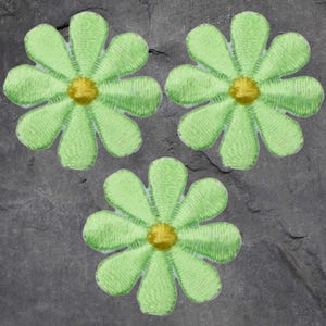 May include: Three embroidered daisy patches. Each patch features a light green petal design with a yellow center. The patches are arranged on a dark gray, textured surface.